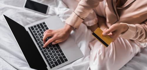 kobieta siedząca na pościeli z laptopem i telefonem sprawdzająca ile kosztuje karta kredytowa