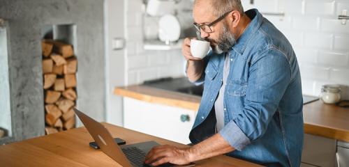 mężczyzna stojący z kawą w kuchni i sprawdzający na laptopie ranking pożyczek 1100 zł