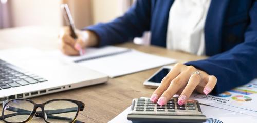 Kobieta w banku liczy wysokość zarobków potrzebną do kredytu 250.000 zł. Na zdjęciu kalkulator, komputer, kobieta w biurze