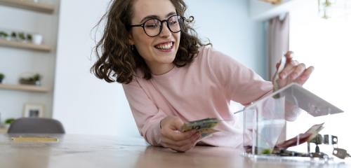 Uśmiechnięta kobieta w okularach wrzucająca banknoty do skarbonki