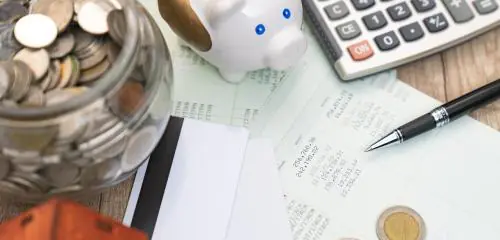 Widok na biurko osoby, która oszczędza: na blacie stoi świnka-skarbonka, słoik z monetami i kartka z podsumowaniem zgromadzonych oszczędności.