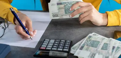 kobieta siedząca przy biurku z plikiem banknotów w dłoni robiąca obliczenia na kartce papieru