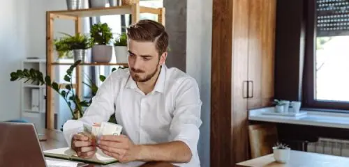 młody mężczyzna siedzący przy stole z banknotami w dłoni i sprawdzający na laptopie ranking pożyczek 1200 zł